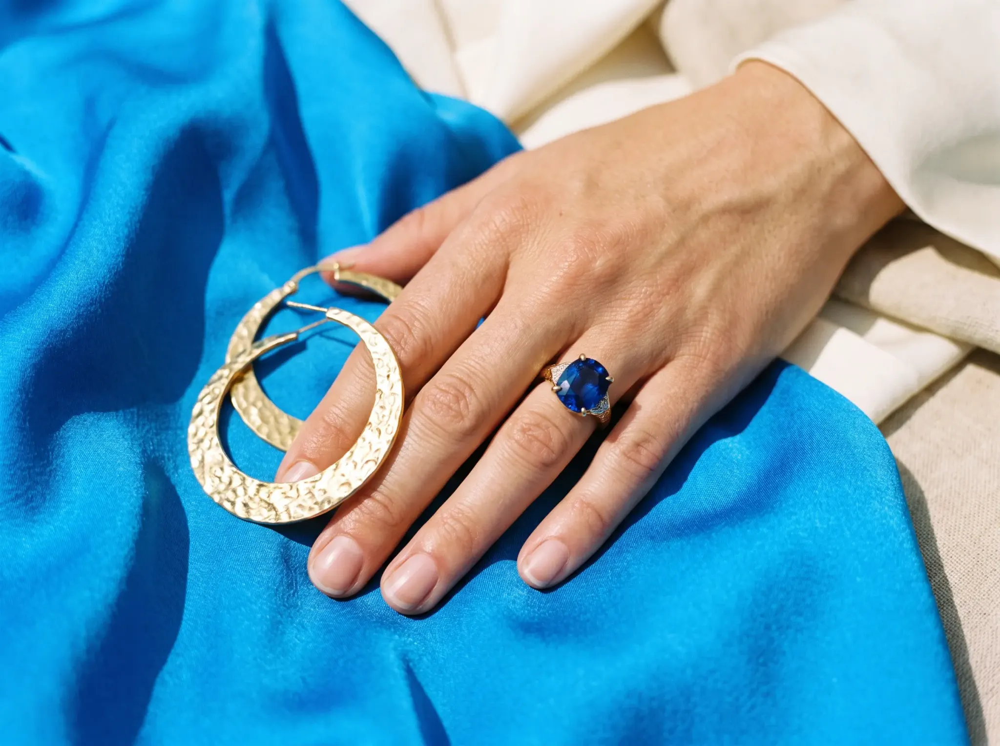 Gold hoops and a sapphire ring on a hand resting on azure silk