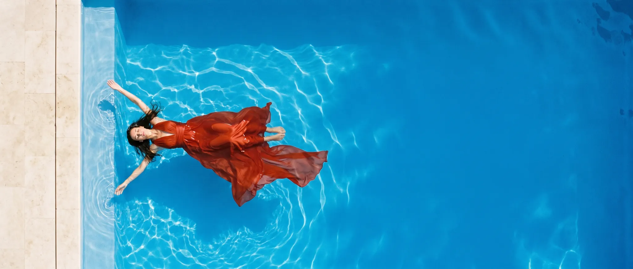 A woman in a vermilion dress floating in an azure pool, seen from above