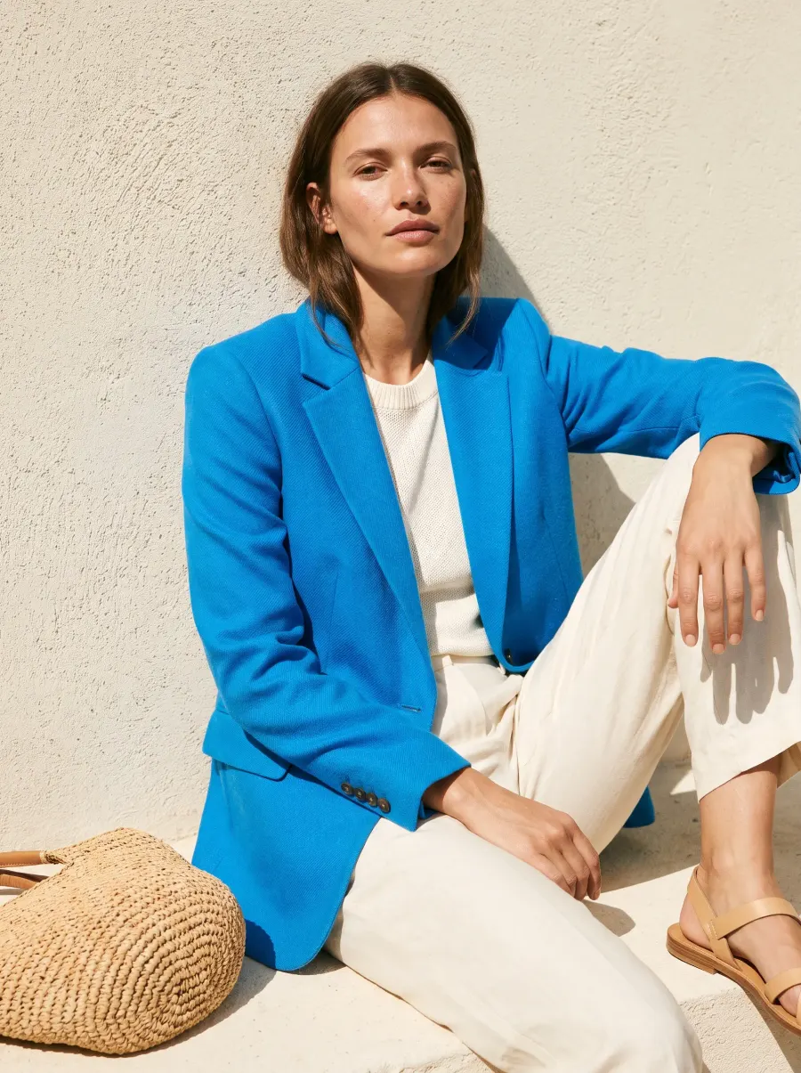A model in an azure blazer leaning on a cream wall in hard sun