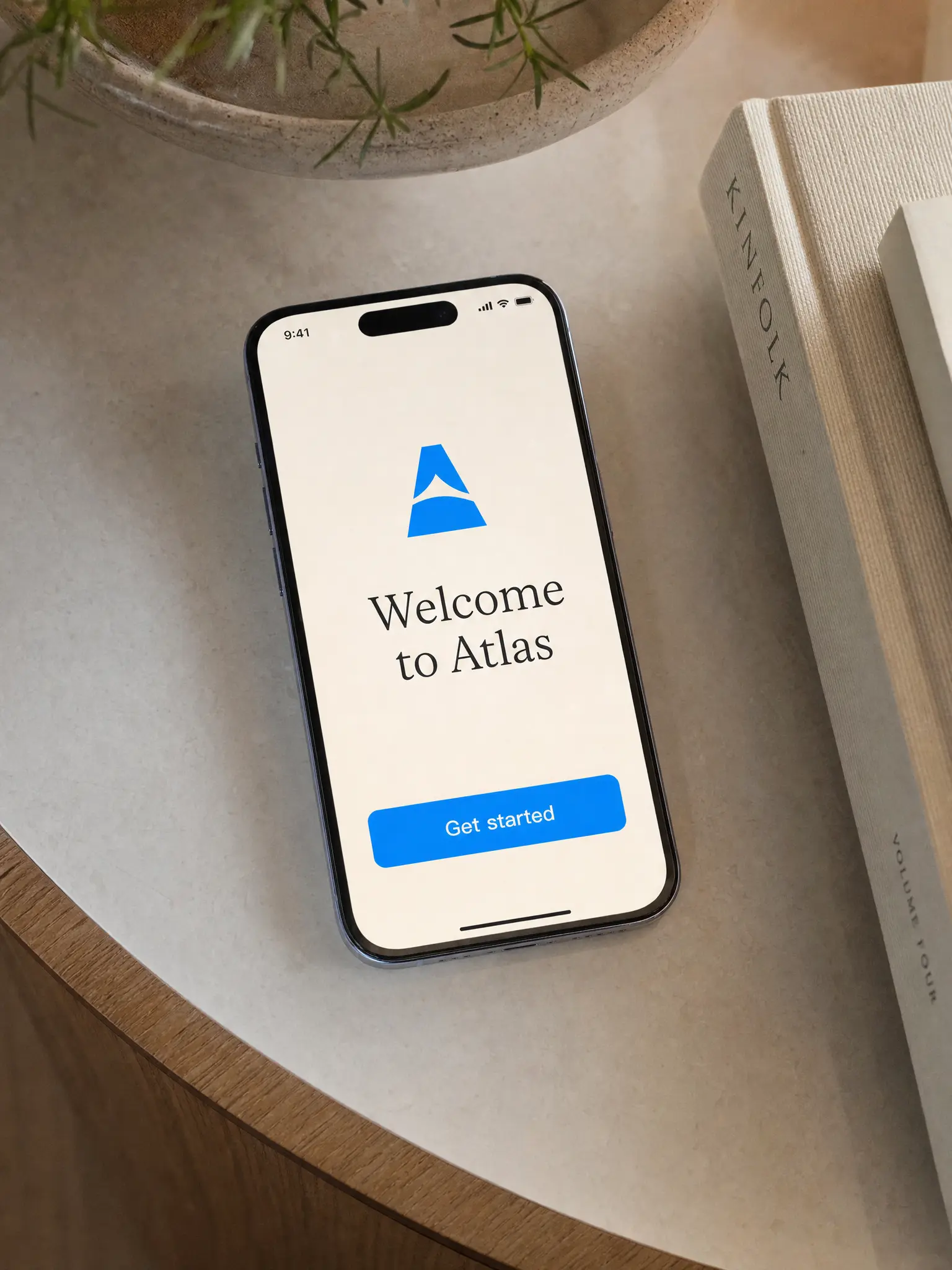 A phone on a desk showing a clean onboarding screen that reads "Welcome to Atlas" with one azure button that reads "Get started"