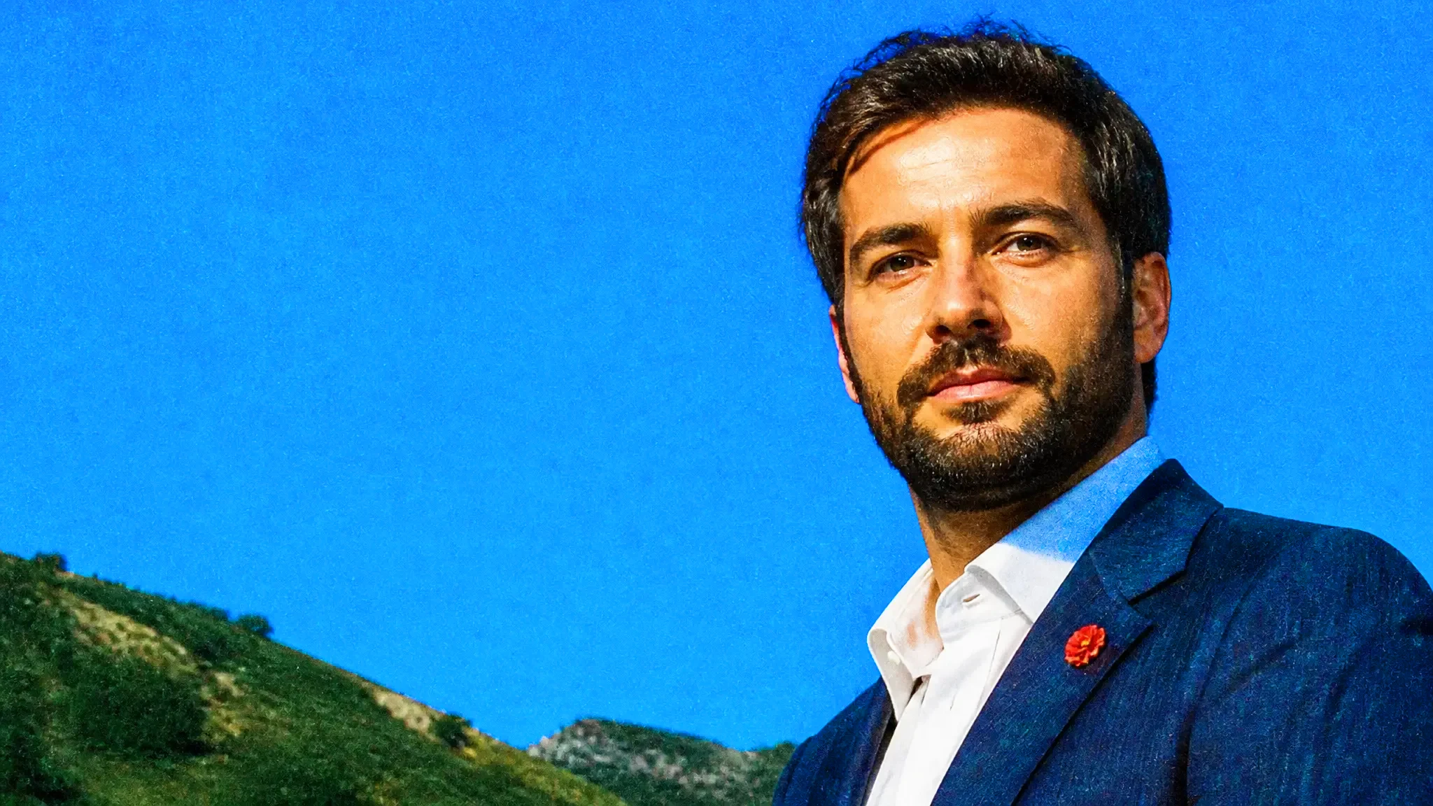A professional headshot of a man in a navy blazer against a blue backdrop