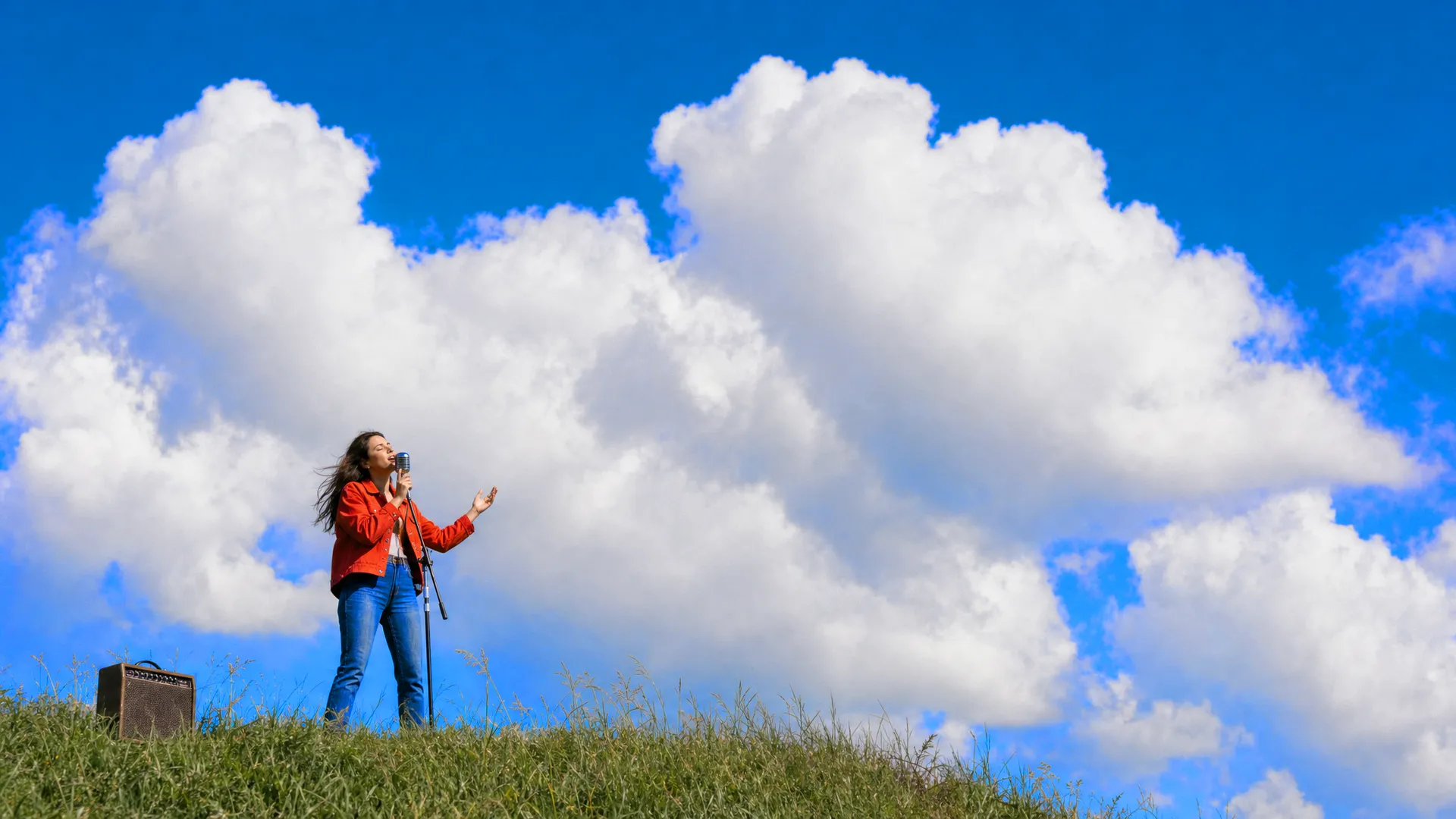 A singer in a red jacket at a vintage microphone on a grassy hilltop, a small amp in the grass beside her
