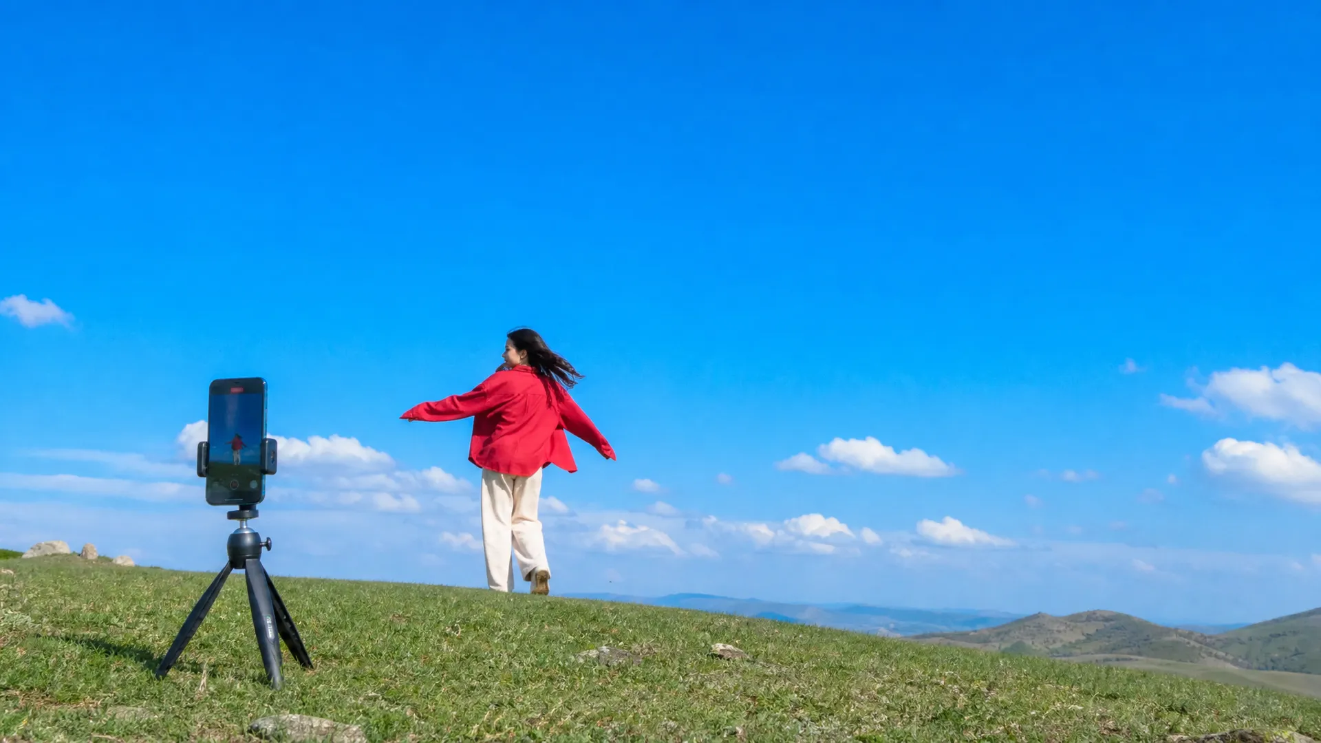 A phone on a small tripod on a hilltop filming a woman in a red jacket mid-twirl