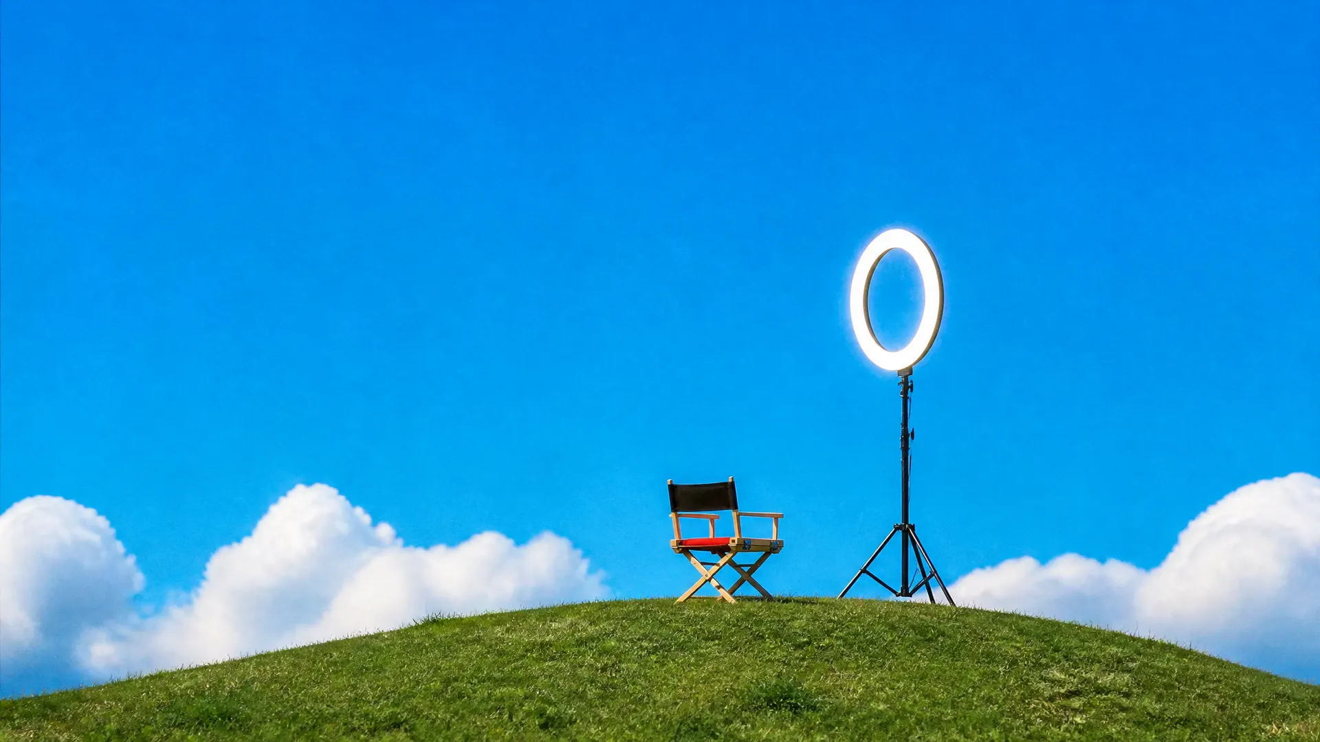 A glowing ring light on a tripod facing an empty director's chair with an orange seat on a grassy hill.