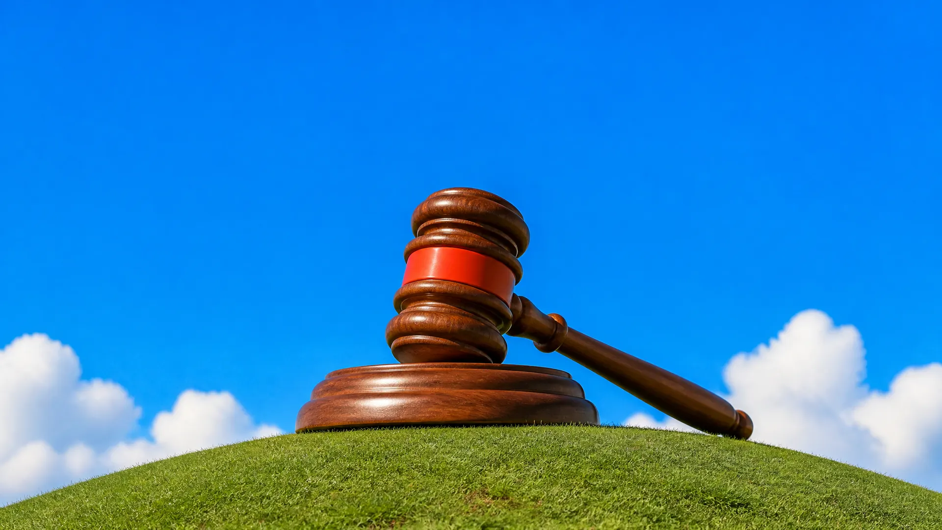 A giant wooden auction gavel with an orange band resting on its block on top of a grassy hill.
