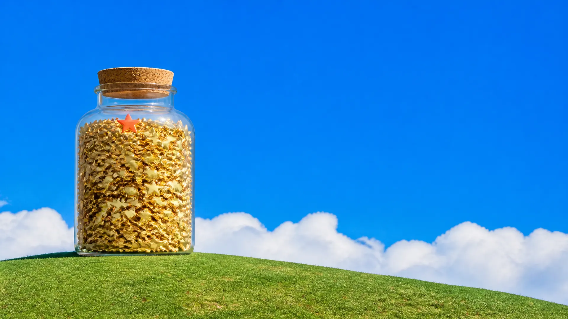 A giant corked glass jar packed with gold stars, one orange star on top, on a grassy hill.