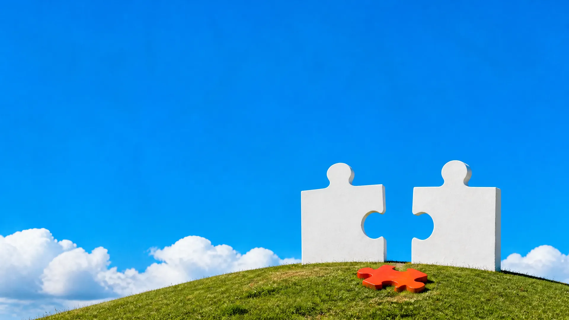 Two white jigsaw pieces standing on a grassy hill with an orange missing piece lying in the gap between them.