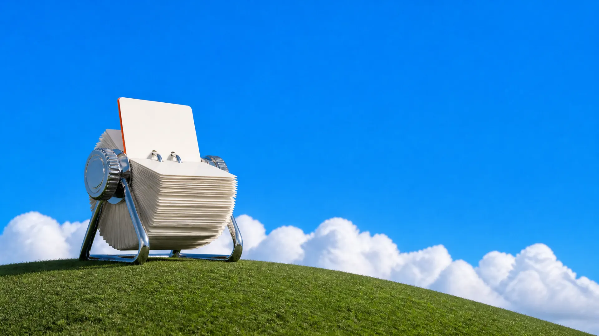 A giant chrome rotary card file with blank index cards, one with an orange edge, on a grassy hill.