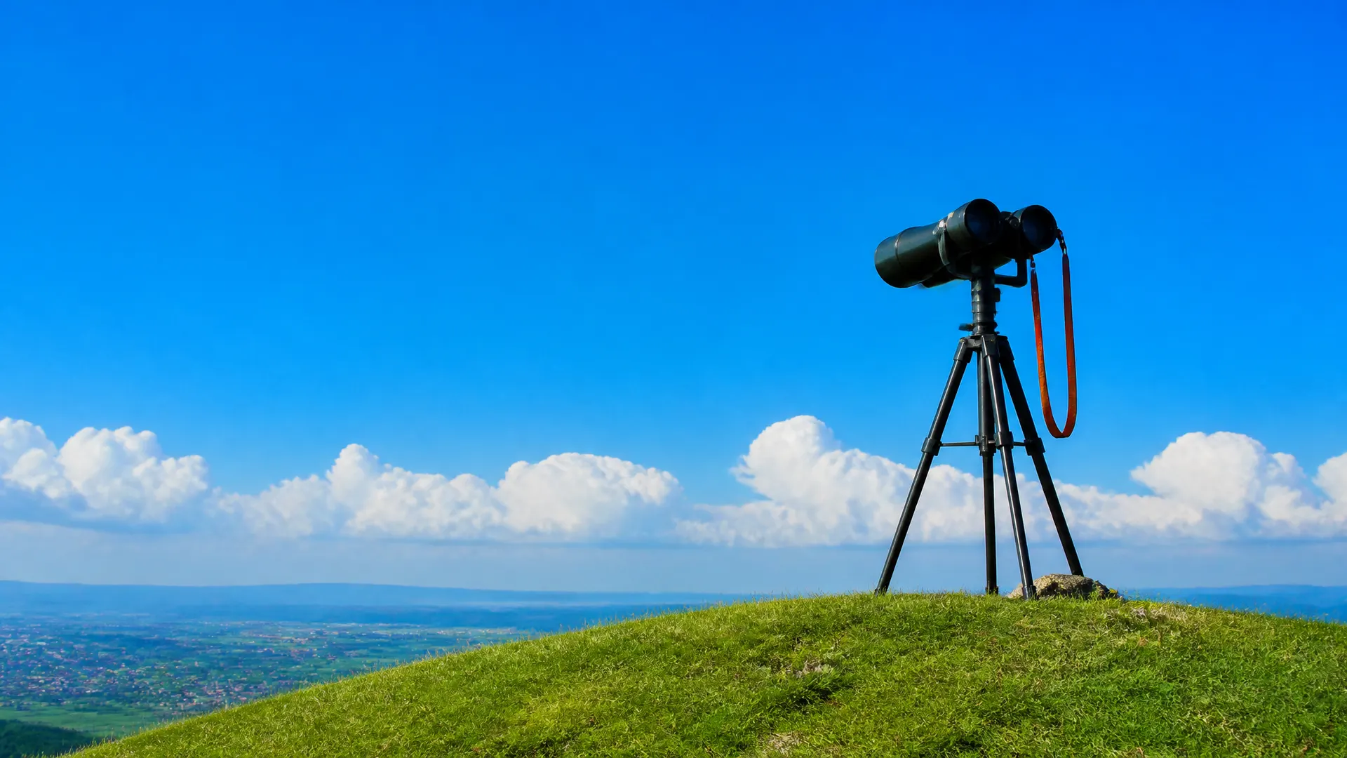 Large binoculars with an orange strap on a tripod at the top of a hill, looking out over a distant valley.