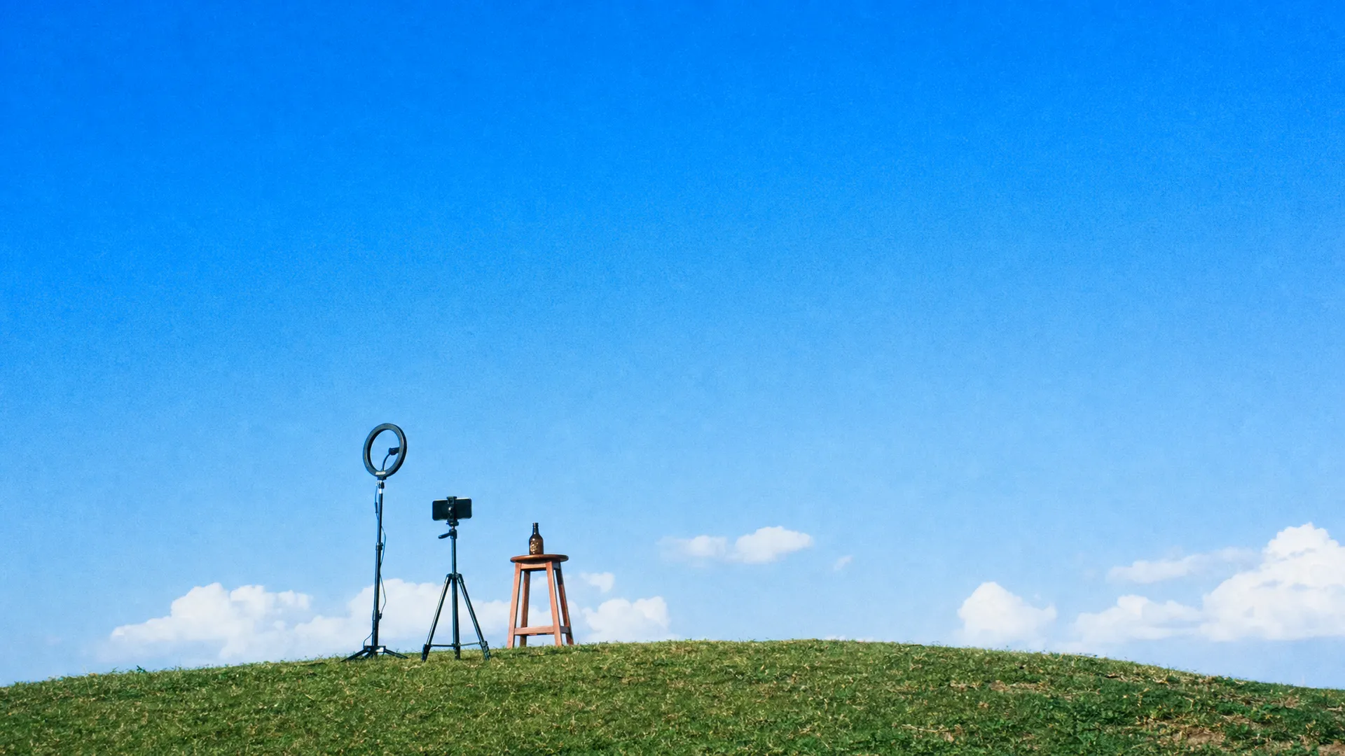 A ring light and a phone on a tripod on a hilltop, filming an amber bottle on a red stool