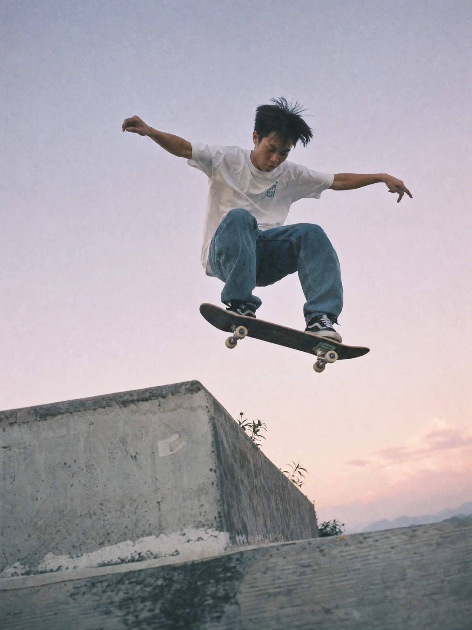 An East Asian skateboarder mid-air against a pastel sky