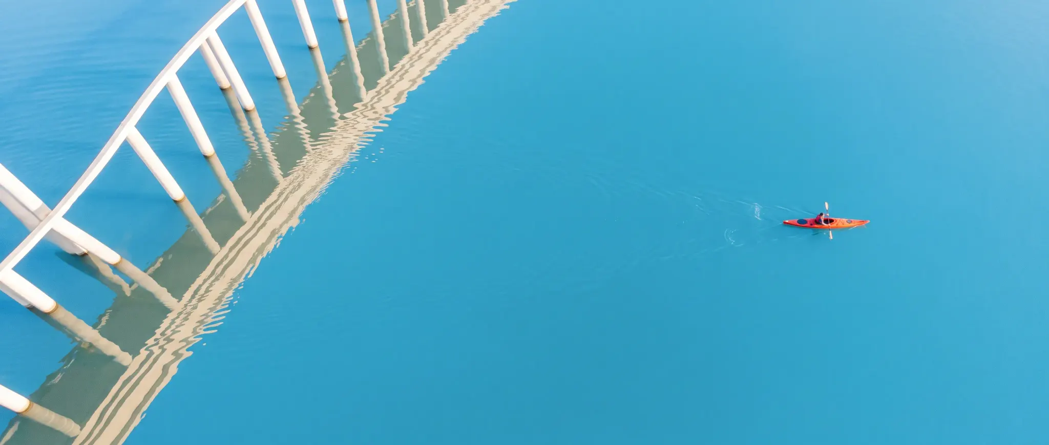 A red kayak crossing blue water with curved architectural reflections