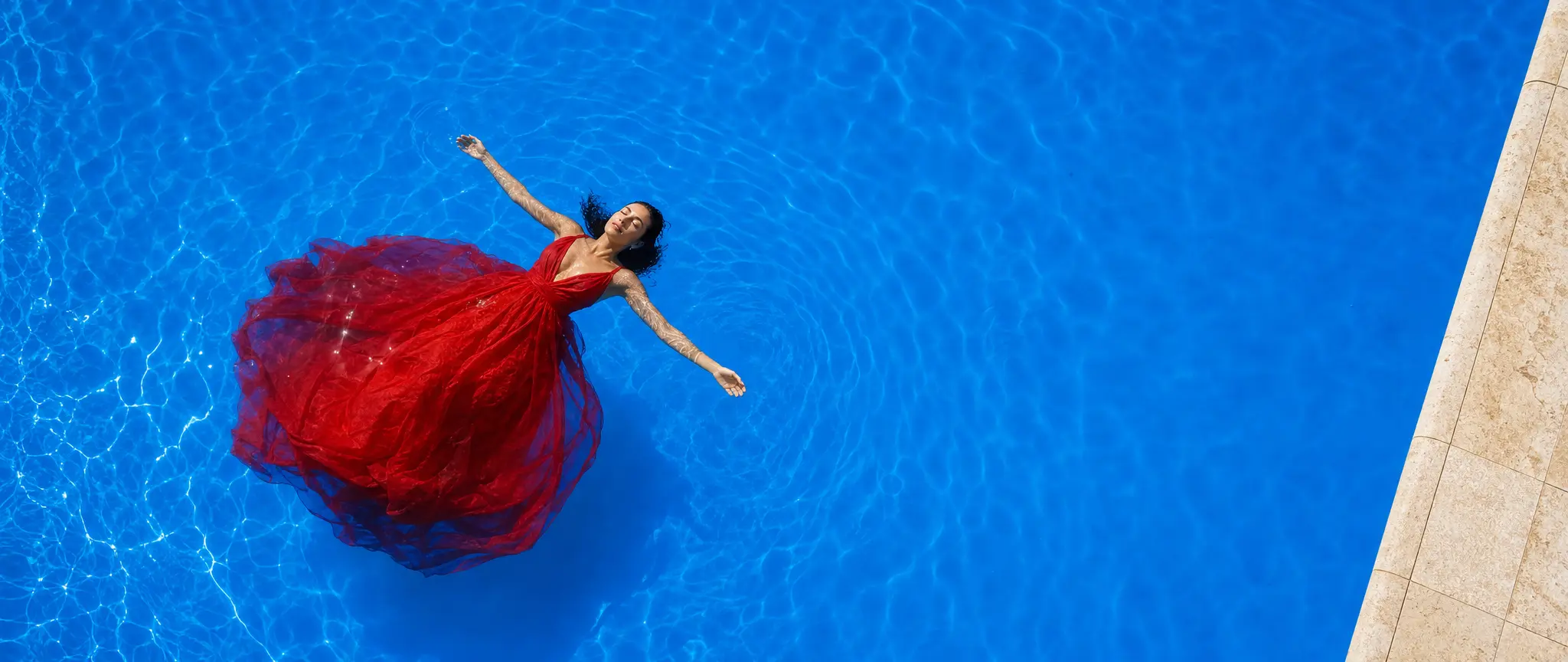 A woman in a vermilion silk dress floating in an azure pool, seen from above